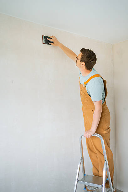 istock Construction worker in brown overalls sanding white wall with sandpaper, balanced on metal ladder, prepping surface for renovation work 2240232481