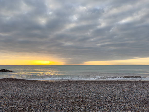 A peaceful morning scene on the pebble beach of Moncofa, Spain. The soft yellow rays of the rising sun break through the clouds, illuminating the calm Mediterranean Sea. The golden light reflects on the wet stones, creating a tranquil and inspiring coastal atmosphere perfect for nature and travel photography