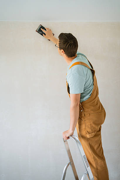 istock Construction worker smoothing white wall with sandpaper, wearing brown overalls and balancing on metal ladder during interior home renovation project 2240233081