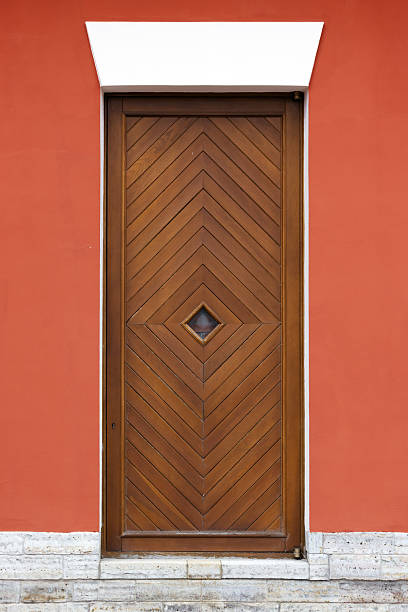 A tall wooden door featuring a chevron pattern sits within a vivid red wall framed by white trim. A small diamond window adds character. Front view, background photo texture