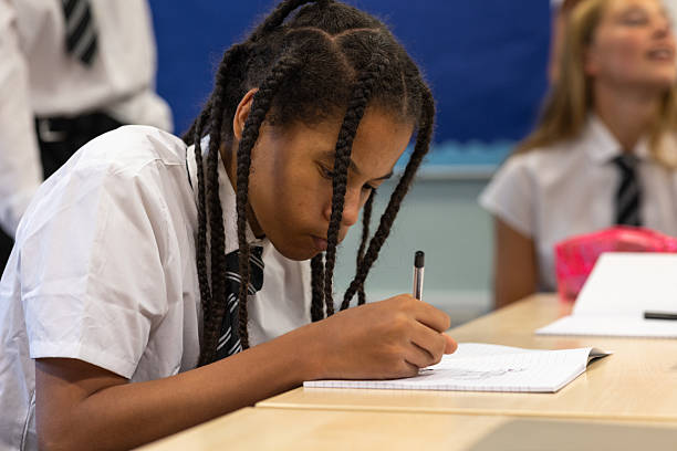 Close-up shot of a teenage boy sitting at a table in the classroom of a school in Newcastle upon Tyne, North East England. He is focusing on the exercise book in front of him, drawing with a pen. There is a teenage girl visible in the background, out of focus, and they are wearing school uniforms.Videos similar to this scenario available.