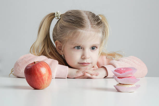 Cute Little Girl at Table Choosing Between Donut and Apple – Healthy Snack and Childhood Concept