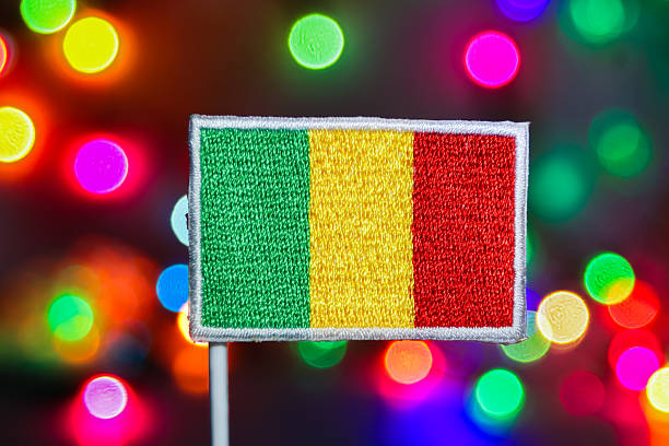 A detailed, embroidered Mali flag featuring the green, yellow, and red vertical tricolor. The flag is sharply focused against a mesmerizing, out-of-focus backdrop of small, multi-colored bokeh lights. This creamy, festive light effect evokes the dazzling spectacle of a New Year's celebration or a major national event. The image is highly versatile for campaigns focusing on West African culture, national pride, and global awareness themes.