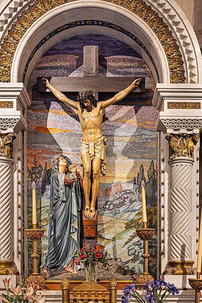 The crypt of Catedral de Santa María la Real de la Almudena, Madrid, Spain, Europe, September 18, 2025: Ornate religious altar inside the crypt of Catedral de Santa María la Real de la Almudena in Madrid, Spain, featuring a central crucifix sculpture of Jesus Christ and a sorrowful statue of the Virgin Mary below. The background includes a vibrant mosaic depicting a radiant sky and hilly landscape, framed by decorative columns and gold accents. Floral arrangements and candles are placed at the base of the altar