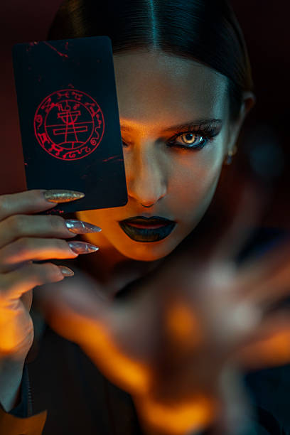 Dark sorceress with glowing eyes and black lipstick holds an occult card, casting a spell with intense, magical energy.
