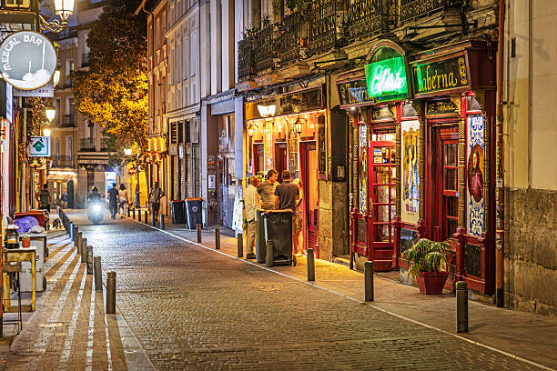 Madrid, Spain, Europe, September 17, 2025: Evening view of a narrow cobblestone street in central Madrid, Spain, lined with small local bars and eateries. The scene features warm street lighting, red-framed windows, and neon signage from a venue named Chicote Taberna, where a small group of people is gathered outside. Bollards separate pedestrian and vehicle zones, and multi-story buildings with balconies frame the street