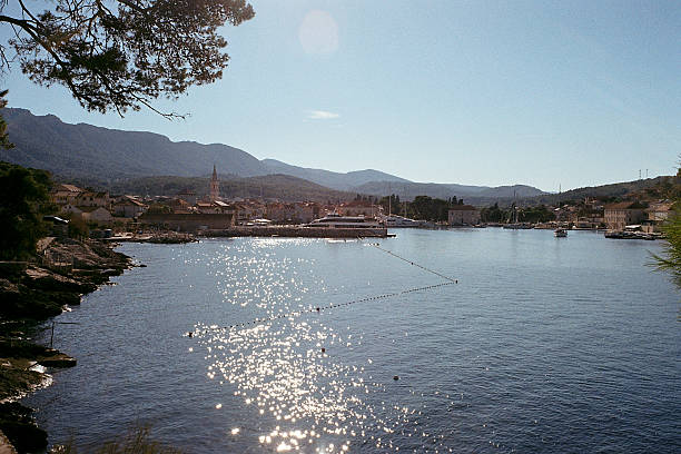 Sunlight sparkling on calm harbor water in a coastal town with moored boats and yachts. Mountain range backdrop and peaceful village buildings along the shoreline. Scenic waterfront view.Shot on film camera