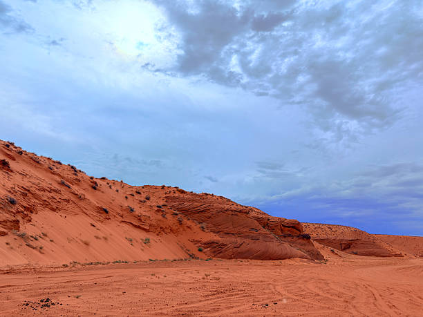Scenic view of striking red sandstone formations and sand dunes beneath a cloudy blue sky in a desert landscape, highlighting natural beauty and geological textures.