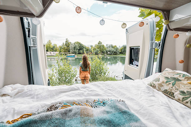 Young woman standing outside camper van by a calm lake surrounded by trees. Cozy van interior with bed and decorative lights. Outdoor relaxation, van life, nature retreat.