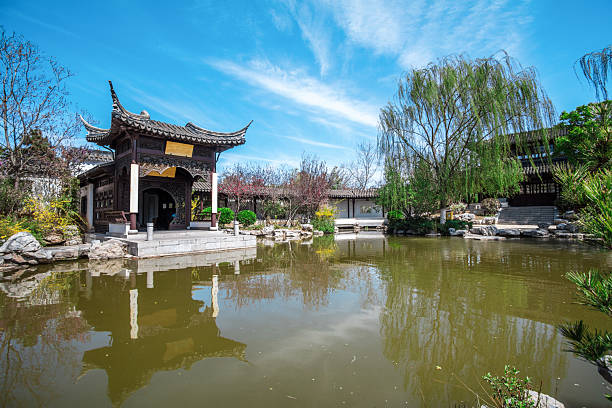 A traditional Chinese pavilion is situated by a calm pond with its reflection visible in the water. The clear blue sky with some clouds, surrounding trees, and garden elements create a serene and picturesque landscape, embodying traditional Chinese architectural beauty and a peaceful natural environment.
