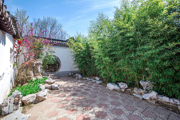 This is a picture of a traditional Chinese garden with a stone path, various plants including bamboo and flowers, an arched door, and some decorative rocks, creating a peaceful and serene outdoor scene