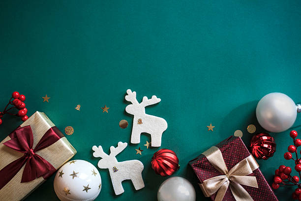 istock Christmas banner design with beige and metallic baubles, glitter confetti, wrapped gift box with red and golden satin ribbon, berries, and felt reindeer on green background. 2240280532