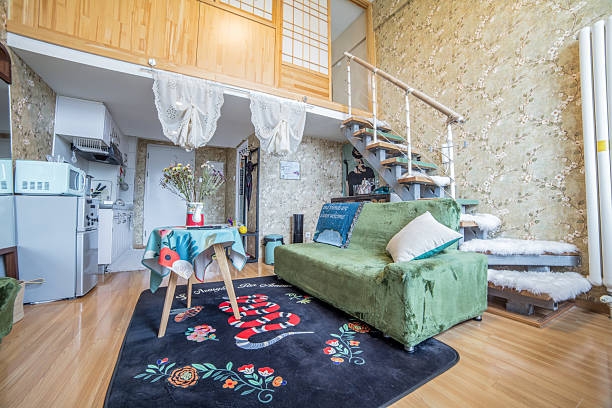 This is a cozy loft apartment with a green sofa, a colorful floral rug, wooden flooring, and a staircase. The interior is decorated in a modern and charming way with a clean kitchen area visible and a warm and inviting living space.