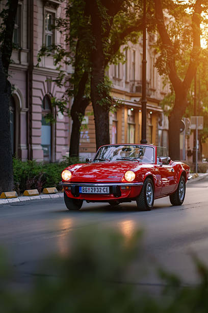 Classic bright red Triumph Spitfire roadster driving down old city street at sunset. Oldtimer travel, iconic vintage style and romantic mood of summer evening. Vertical. Subotica, Serbia, 05 July 2025