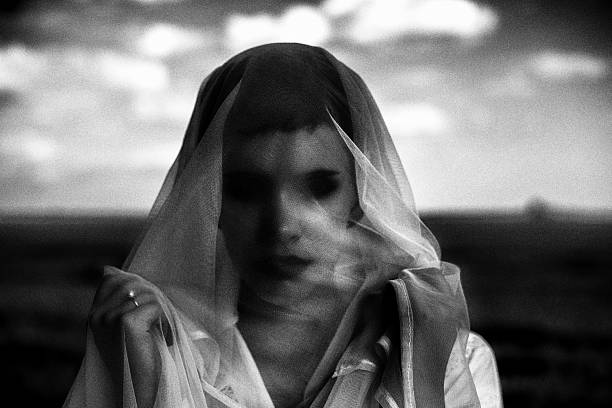 Dark spooky portrait of the young bride under veil standing in the field in black and white.