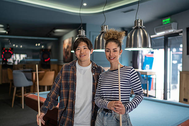 Young adult couple standing by a pool table, holding cue sticks and smiling at the camera, enjoying their leisure time together in a vibrant game room