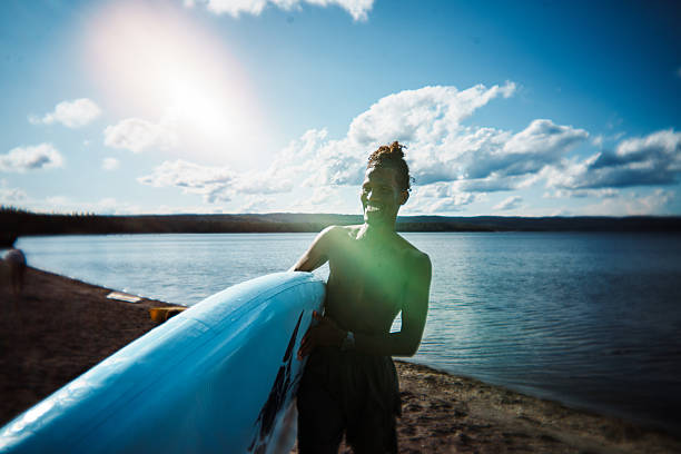 A smiling young Black man carries a paddle board along the sandy shore of Yellowstone Lake in Yellowstone National Park, Wyoming, USA, while celebrating the Fourth of July and camping with a group of friends.