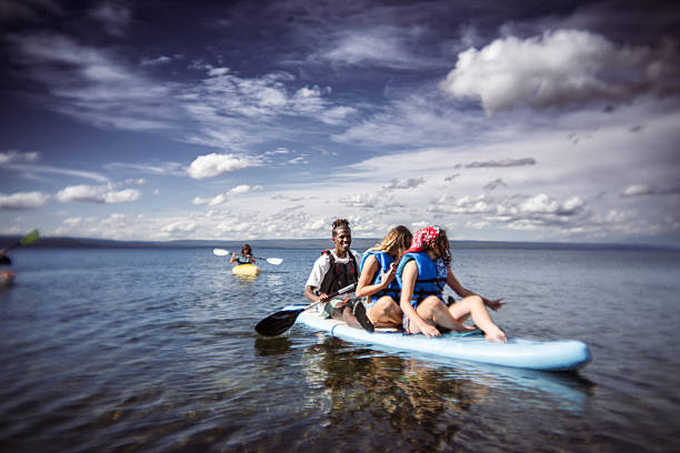 A young man and two young women wearing life jackets laugh and enjoy paddle boarding together on the calm waters of Yellowstone Lake in Yellowstone National Park, Wyoming, USA, on a sunny summer day.