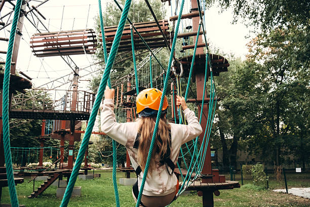Young child wearing safety gear makes their way across a challenging ropes course in an adventure park. The image captures focus and determination during this outdoor activity amid lush surroundings.