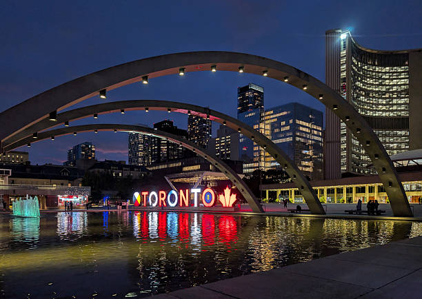Toronto, Canada - August 26, 2025: Looking northwest from Nathan Phillips Square. The illuminated TORONTO sign reflects in the water fountain by City Hall. Summer twilight in the Yonge-Bay Corridor downtown.