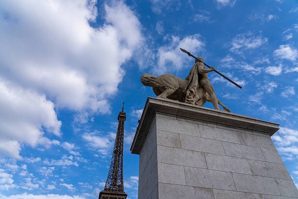 A statue of a centaur and a man holding a spear stands against a blue sky with clouds, with the Eiffel Tower visible in the background.