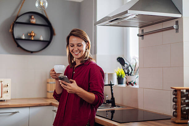 istock Young woman smiling while holding a cup of coffee and using smartphone in the kitchen 2240335559