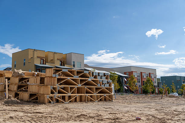 Multi-story residential buildings under construction with exposed wooden framing structures in foreground. Unfinished housing development site with partially completed buildings, sandy terrain, and clear blue sky background. Modern urban expansion project with structural framework visible.