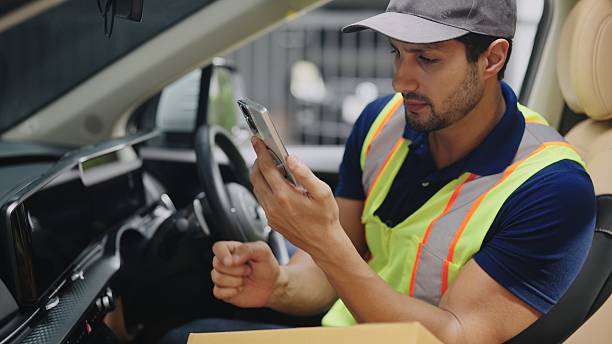 Delivery man  wearing blue polo checking his smartphone for cardboard box package tracking details sitting in van ,courier logistics and courier service process