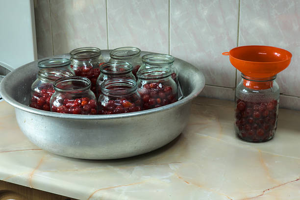 Eight glass jars filled with fresh cherries sit on a kitchen countertop. One jar is being filled with cherries through a funnel, creating a calm and productive domestic atmosphere.