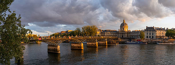 People stroll across the Pont des Arts in Paris at sunset, enjoying the view of the Seine River and the Institut de France.