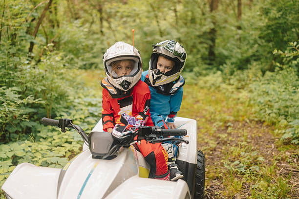 Two young boys wearing helmets and colorful outfits ride an ATV on a forest trail surrounded by lush greenery on a sunny day.