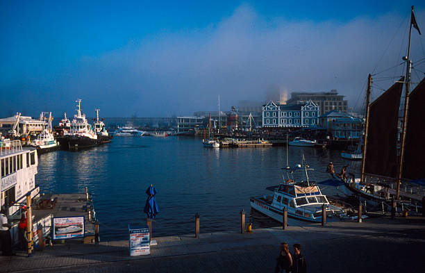 Sunlit Cape Town harbor with colorful boats, calm green water, and a scenic backdrop of Table Mountain and the Twelve Apostles. A vibrant coastal scene perfect for travel and lifestyle use.