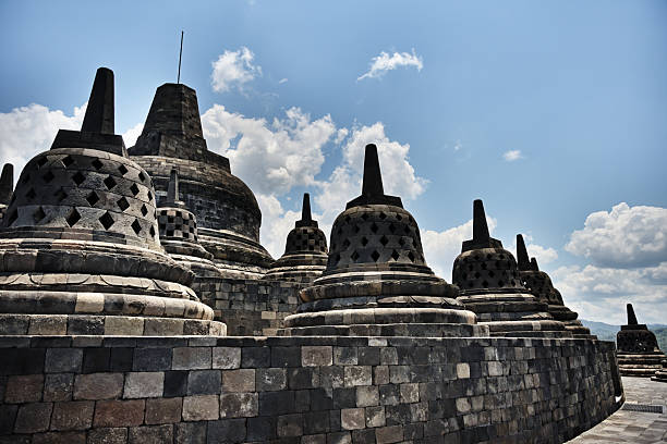 In the heart of the island of Java, the Borobudur temple unfolds like a giant mandala carved from volcanic rock. On its upper terraces, a paved walkway winds between openwork stupas, each housing a Buddha statue turned toward spiritual awakening. The gaze is lost in the perfect geometry of these stone bells, aligned with age-old precision, while distant mountains appear on the horizon under a pure blue sky.A symbol of inner quest, each step along this circular path evokes a journey toward knowledge and serenity. The balance between the architectural rigor and the gentle surrounding landscape creates an almost timeless atmosphere.Borobudur, a UNESCO World Heritage Site, remains one of the greatest Buddhist sanctuaries in the world, a place where faith, nature, and art meet in perfect harmony. This panorama invites contemplation, slowness and inner peace, like the Buddhism it has embodied for over a millennium.