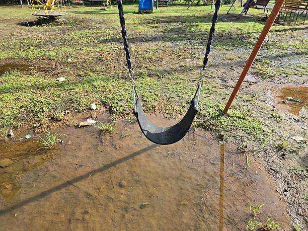Closeup of an empty playground swing hanging above a muddy puddle after rainfall. Wet ground with grass and reflections in the water under bright sunlight, symbolizing childhood memories, calmness, and monsoon atmosphere.