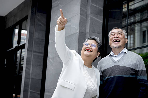 istock Happy senior Asian couple pointing somewhere while standing outside the building. Smiling mature elderly lover standing outdoor on the city walkway. Retired people spending time together outdoors. 2240406328