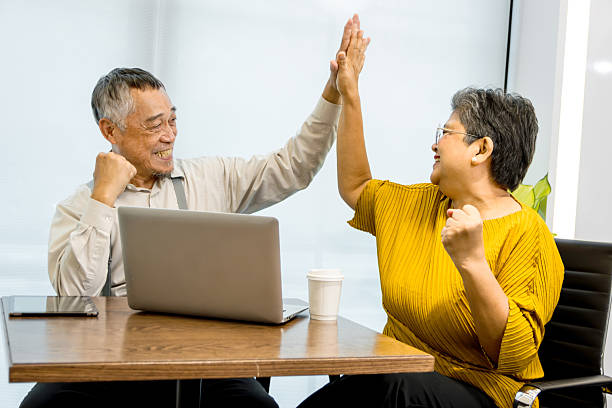 istock Two senior businesspeople work with laptop together and make high five hand touch to celebrate of success. Happy elderly Asian businesswoman and mature manager touch hands with hi five in office. 2240408257