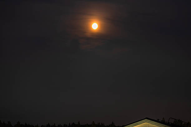 Bright full moon glowing through thin clouds in night sky above dark forest and roof edge villa. Sweden.