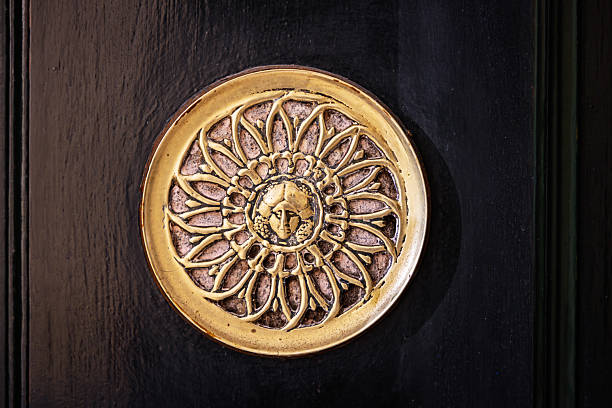 Close-up of a decorative brass grille mounted on a dark green door in Madrid, Spain. The circular ornament features an intricate radial design with petal-like elements and a sculpted central face, possibly mythological. Historically used as a secure voice portal, the grille allowed residents to speak with visitors without opening the door. The craftsmanship reflects traditional Spanish architectural detailing and functional design, suitable for themes of heritage, urban security, and cultural preservation.