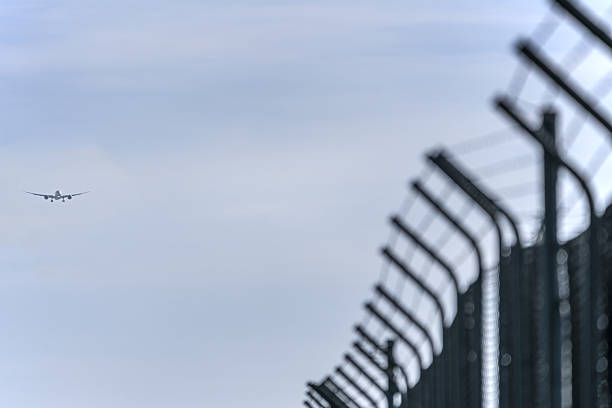A plane approaching the airport to land on a runway whose perimeter is protected by a metal fence with concertina.