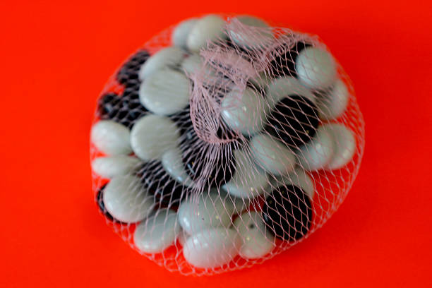 ​A white mesh bag is filled with smooth decorative stones alternating between milky-white and glossy black colors. The object is shot close-up against a bright orange background, creating a strong color contrast.