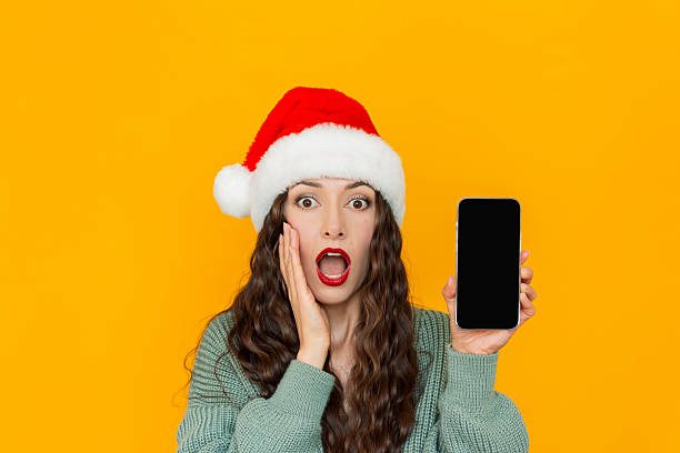 istock Young woman wearing Santa hat holding smartphone with empty screen and looking at camera with surprise 2240443693