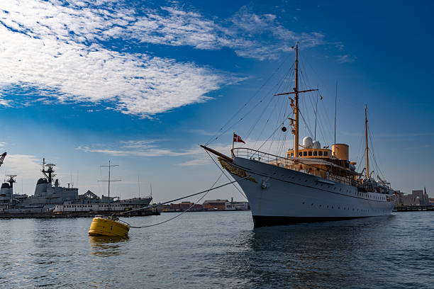 Copenhagen, Denmark-August 14, 2025: Classic royal yacht anchored at pier, Danish flag waving, surrounded by calm water, blue sky, light clouds, naval ships nearby.