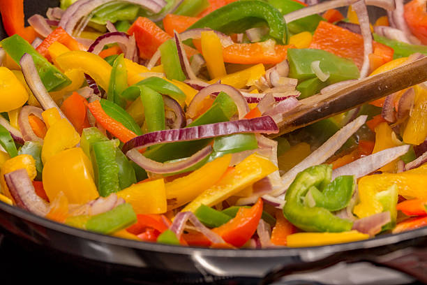 Close up of an identifiable chef stirring chopped red peppers, green peppers, yellow peppers and red onions with a wooden spoon