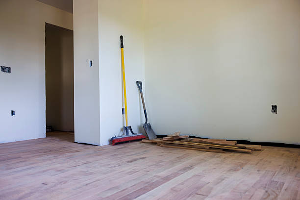 Construction tools including broom, shovel and wooden boards sit against a white wall in an empty room undergoing renovation. The space features newly installed light wood flooring, unfinished walls with exposed electrical outlets, and a doorway leading to another area. The room is in mid-construction phase with no furniture or finished elements present. Wide shot with deep focus showing the entire empty space.