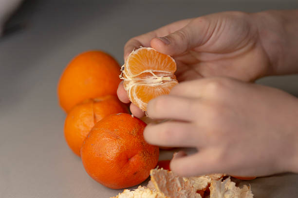A childs small hand is carefully peeling a bright mandarin orange, with several other oranges nearby on a table