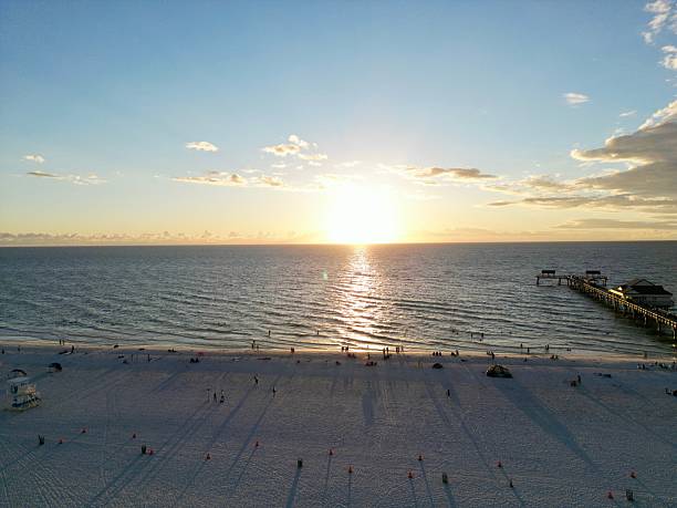 Aerial view of a serene beach at sunset with a long pier extending into the ocean, capturing the golden hues and calm waters.