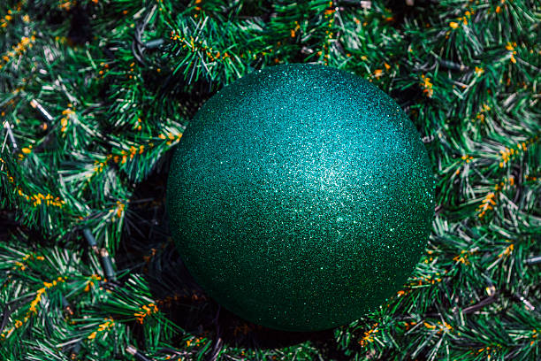 Shimmering green sphere glitter ornament is nestled in the branches of a dark green Christmas tree with small fairy lights. Ornament positioned centrally among the synthetic pine needles