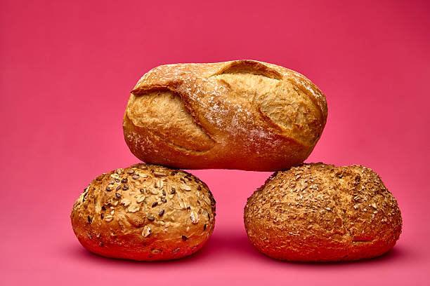 Horizontal composition of a small bread loaf placed over two round seeded buns against pink background. Modern minimalist food still life ideal for bakery branding.