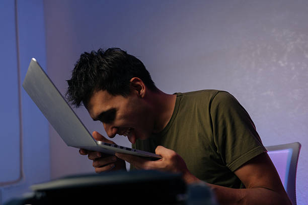 Young programmer male showing extreme frustration by licking laptop after software crash, expressing desperation and anger in humorous way. Frustrated software developer licking laptop keyboard.