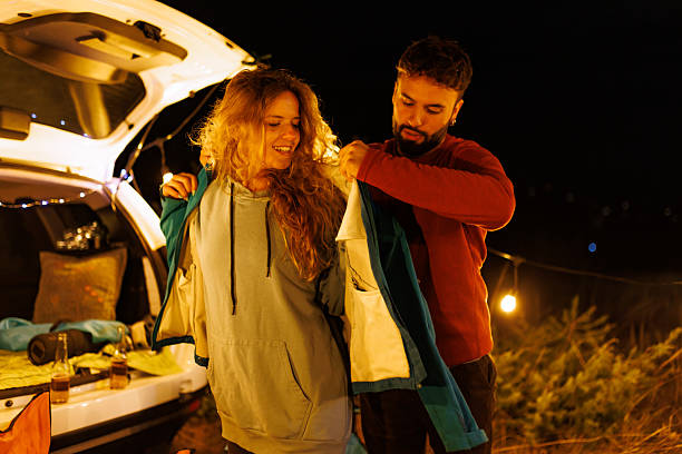 istock Couple sharing a warm moment outdoors while putting on jackets at a cozy campsite during a chilly night, surrounded by nature and soft lighting 2240494441
